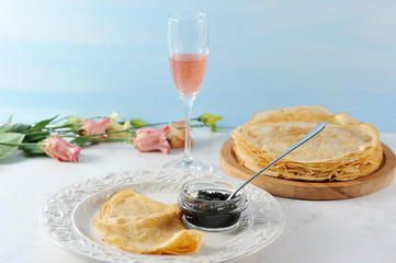 Thin pancakes on a wooden tray. Next to the plate with pancakes and black caviar. In a cup of caviar a teaspoon. In the background, pink flowers Eustoma and a glass of pink champagne.  
