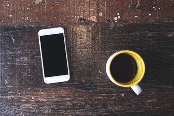 Top view mockup image of a white mobile phone with blank black desktop screen with coffee cup on vintage wooden table