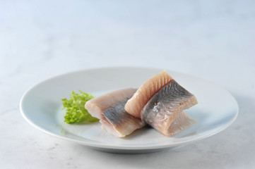 Fillet of Atlantic herring on a white plate. The fish is decorated with a leaf of lettuce. Light marble background. Close-up. Macro photography.