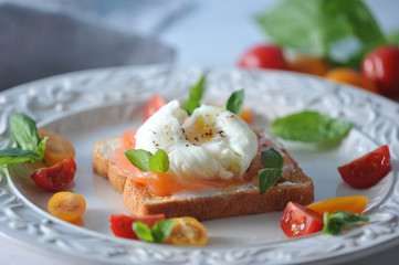 Slices of salmon fillet of weak salt on white bread toast. On top of the egg is poached. The toast is decorated with multi-colored mini tomatoes  and basil leaves. Light background. Close-up. Macro.