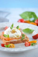 Toast with salted salmon and egg poached on a plate. The dish is decorated with multi-colored cherry tomatoes and basil leaves. Light background. Close-up. Vertical orientation of the frame.