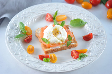 Slices of salmon fillet of weak salt on white bread toast. On top of the egg is poached. The toast is decorated with multi-colored mini tomatoes  and basil leaves. Light background. Close-up. Macro.