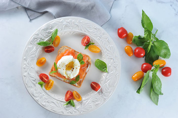 Toast with salted salmon and egg poached on a plate. The dish is decorated with multi-colored cherry tomatoes and basil leaves. Light background. Close-up. View from above. 