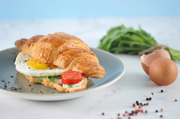 Crispy croissant with arugula, fried egg, slice of tomato. In the frame, a bundle of arugula and eggshell. Light background. Close-up.