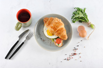 Croissant with arugula, fried egg, slice of tomato. In the frame is a cup of coffee, a bunch of arugula and cutlery. Light background. View from above.