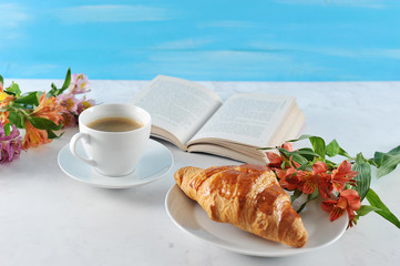 A cup of coffee and a crispy croissant. On the plate with croissant flowers. In the frame an open book. Next to the book is a bouquet of flowers. Light background. Close-up. 