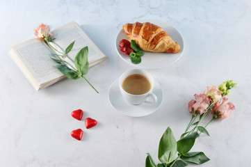 A cup of coffee and a crispy croissant. Around the candy in the shape of the heart. In the frame an open book, on the pages of the book lies a flower. Light background.. View from above.