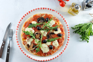 Pizza on the platter. The structure of the filling includes olives, mushrooms, salami, arugula, mozzarella, tomatoes. In the frame cutlery and a bunch of arugula. Close-up. View from above.