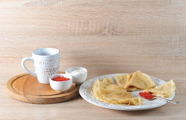 Pancakes on a plate and a teaspoon with caviar. Next to the wooden tray. On the tray there is a cup of tea, bowls with red caviar and sour cream. Light wooden background. Close-up.