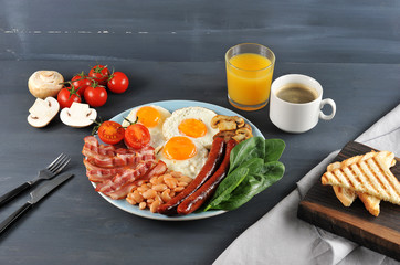 English breakfast - brunch. On a plate fried bacon, sausages, fried fresh tomatoes and champignons, white beans in tomato sauce, toast, fried eggs. In the frame, a glass of orange juice and coffee.