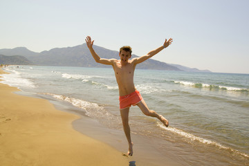Funny young man jumps and laughs on the wide beach. Waves with foam comes to the shore. Athletic guy in pink swimming trunks. Mountains and sun on background. hipster holiday summer relaxing lifestyle