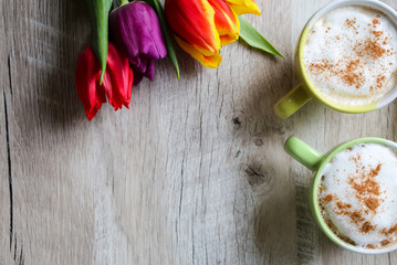 Tulips on wooden background with two cups of coffee. Invitation postcard for mother's day or international women's day. Punchy pastels. Cappuccino with foam cinnamon and flowers.