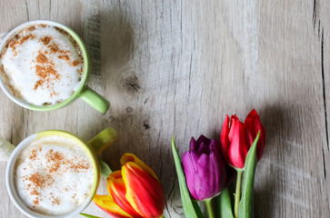 Tulips on wooden background with two cups of coffee. Invitation postcard for mother's day or international women's day. Punchy pastels. Cappuccino with foam cinnamon and flowers.