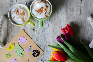 Tulips on wooden with two cups of coffee. Invitation postcard for mother's day or international women's day. Spring paper birds on craft paper envelope. Handmade origami. Punchy pastels. Cappuccino.