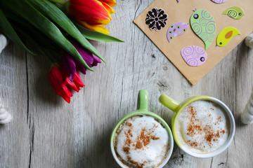 Tulips on wooden with two cups of coffee. Invitation postcard for mother's day or international women's day. Spring paper birds on craft paper envelope. Handmade origami. Punchy pastels. Cappuccino.