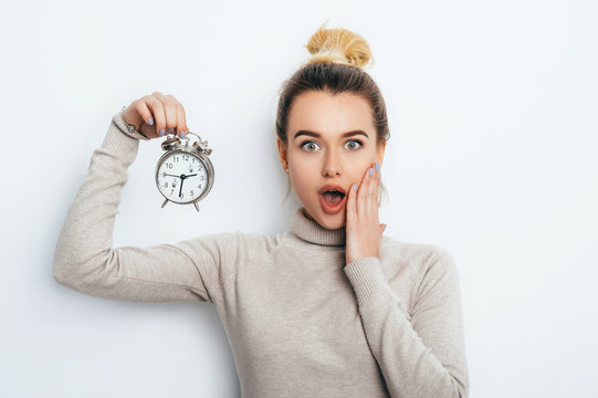 Portrait of beautiful young cheerful blonde woman with hair bun in sweater holding alarm clock and show emotions while standing over solated white background. Lifestyle Fashion Beauty Business Student