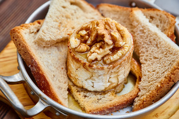 Snack from oven-baked goat cheese with walnuts and roasted homemade bread in an aluminum saucepan on a round light wooden cutting board on a brown wooden table. With cheese balls and sauce.