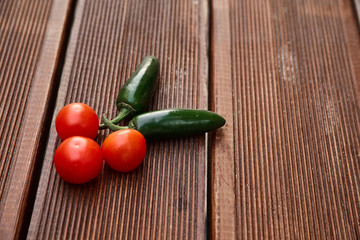 A conceptual composition of red cherry tomatoes and hot green chili peppers on dark brown wooden boards with a deep texture. Stylish minimalism. Photographed in natural daylight.