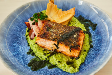 Large juicy roasted fatty piece of salmon steak on a cushion of vegetable puree, decorated with radish, parsley, cilantro and baked dough. Fish dish in a beautiful blue plate on a white wooden table.