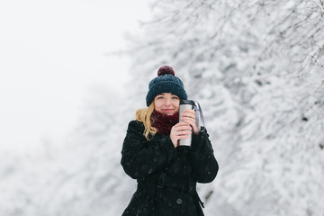 Fototapeta na wymiar Young caucasian blonde girl in black coat and red knitted scarf walks in winter park with thermos bottle