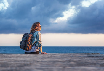 portrait of a beautiful happy young woman sitting on a wooden pier on the background of the sea and a beautiful sunset and admiring the scenery