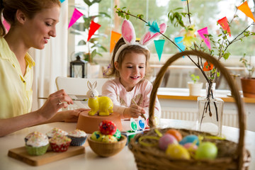 A mother and daughter celebrating Easter, painting bunny with brush in yellow color. Happy family smiling and laughing. Cute little girl in bunny ears preparing the holiday.