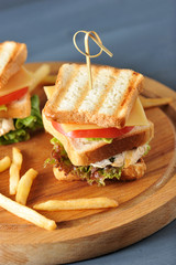 On a wooden tray there are mini sandwiches made of fried white bread and french fries. The filling of the sandwich consists of grilled chicken, salad, cheese, slice of tomato. Dark background. Closeup