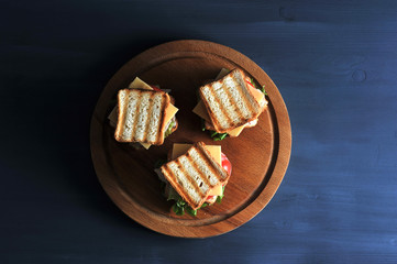 On a wooden tray there are three mini sandwiches made of white bread. The filling of the sandwich consists of grilled chicken, salad, cheese, slice of tomato. Dark background. View from above.