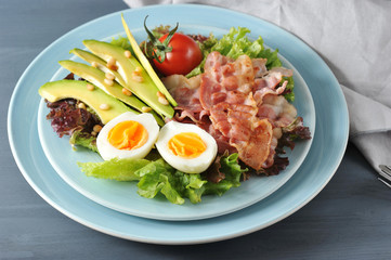 Salad with avocado and bacon. In a deep plate lettuce leaves, hard-boiled egg, fried bacon, avocado slices, tomato, pine nuts. In the picture, a napkin.  Dark background. Close-up. Macro photography.