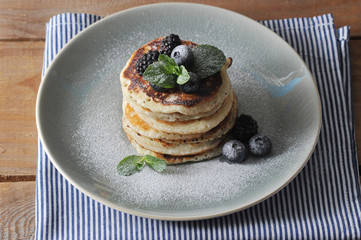 Pancakes on a blue plate. Pancakes are decorated with blueberries, blackberries, mint and sprinkled with powdered sugar. Under the plate is a napkin. Wooden background. Close-up.