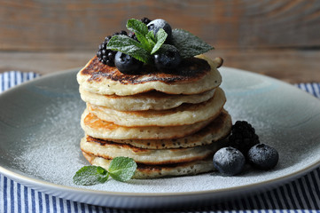 Pancakes on a blue plate. Pancakes are decorated with blueberries, blackberries, mint. The dish is sprinkled with sugar powder and decorated with berries and mint. Under the plate is a napkin. Closeup