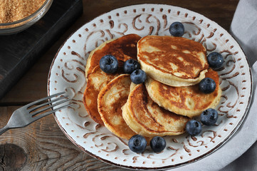 Small pancakes in a plate. Appetizing pancakes are decorated with blueberries. Next fork. Rough wooden background. Close-up. Macro photography.