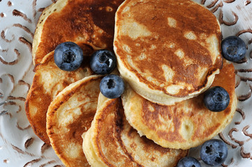 Small pancakes in a plate. Appetizing pancakes are decorated with blueberries. Next fork. Rough wooden background. View from above. Close-up. Macro photography.