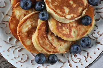 Small pancakes in a plate. Appetizing pancakes are decorated with blueberries. In the frame is part of the plate. Rough wooden background. View from above. Close-up. Macro photography.