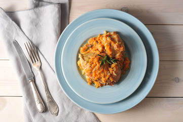 Rabbit is cooked with rosemary and carrots. The dish is decorated with a branch of rosemary. In the frame cutlery and napkin. Light wooden background. View from above. Close-up. 