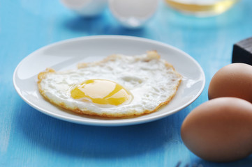 Fried eggs in a white plate. In the frame, a few eggs are eggs in the shell. Blue wood background. Close-up.  Macro photography.