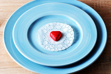 Two blue plates one on the other. A white paper napkin is on the plate. On a napkin is a red candy in the shape of a heart. There are cutlery nearby.  Light wooden background. Close-up.  Macro.