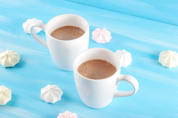 Two cups of hot cocoa. A fragrant drink energizes and creates a mood. Round the cups are arranged meringues. Blue wood background. Close-up.