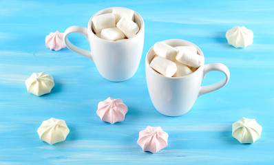 Two cups of hot cocoa with marshmelow. A fragrant drink energizes and creates a mood. Round the cups are arranged meringues. Blue wood background. Close-up.
