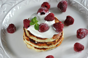 Pancakes are laid one on top of another on a white plate. On top of the pancakes are sour cream and decorated with raspberries and mint leaves. Close-up. Macro photography.