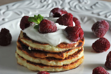 Pancakes on a plate are laid one on another. On top of the pancakes are sour cream and decorated with raspberries and mint. Wooden background. Close-up. Macro photography.