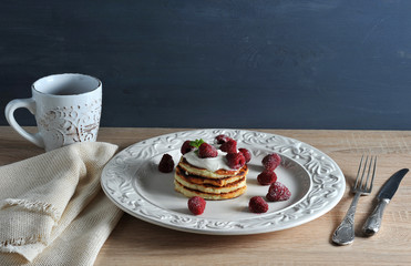 Pancakes on a plate lie one on the other. From above pancakes are decorated with sour cream, raspberries and mint. Next cutlery, napkin, a cup of coffee. Wooden background. Close-up.