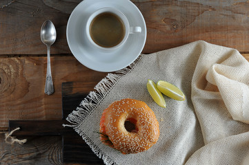 Beagle with salmon, soft cheese and dill on a cloth napkin. There are two slices of lemon. In the frame is a cup with black coffee and a teaspoon. Close-up. View from above.