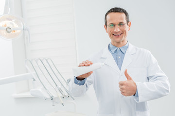 Naklejka na ściany i meble Dentist showing blank toothpaste tube and thumb up in modern dental clinic