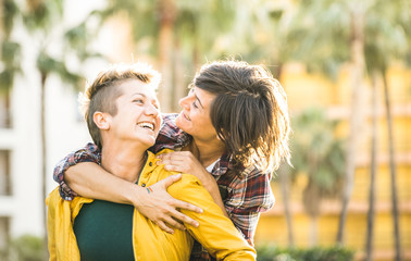 Naklejka premium Happy playful girlfriends in love sharing time together at travel trip on piggyback hug - Women friendship concept with girls couple having fun on fashion clothes outdoors - Bright warm sunset filter