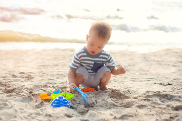 Cute little Asian 18 months, 1 years old toddler boy sitting & playing children's beach toys  on beautiful sandy tropical sunset beach, Family travel, water outdoor activity on beach vacation concept