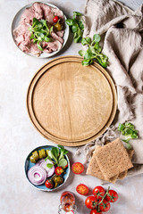 Ingredients for making sandwiches. Sliced beef meat, rye whole graine bread, green salad, tomatoes, pickled cucumber, onion on plates, empty wooden tray over grey texture background. Top view, space.