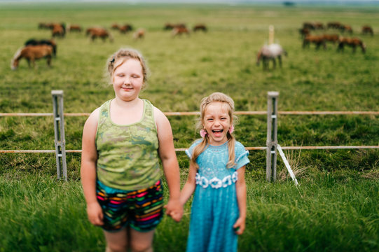 Tilt shift blurred expressive mood potrtrait of two little sisters resting outdoor in summer together. Young girls with emotional faces. Chilhood in village. Funny kids near grazing horses field.