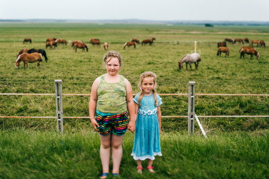 Tilt shift blurred expressive mood potrtrait of two little sisters resting outdoor in summer together. Young girls with emotional faces. Chilhood in village. Funny kids near grazing horses field.
