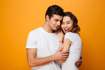 Portrait of handsome man in white t-shirt hugging his girlfriend while standing on camera, isolated over yellow background